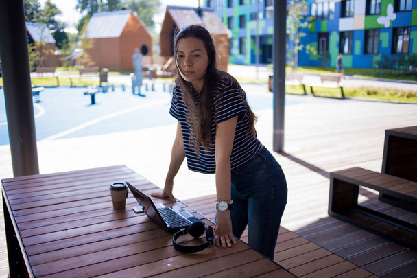 Serious brunette girl, stands near the wooden table with gadgets, headphones, laptop, smartphone. - Fotografie, Obrázek