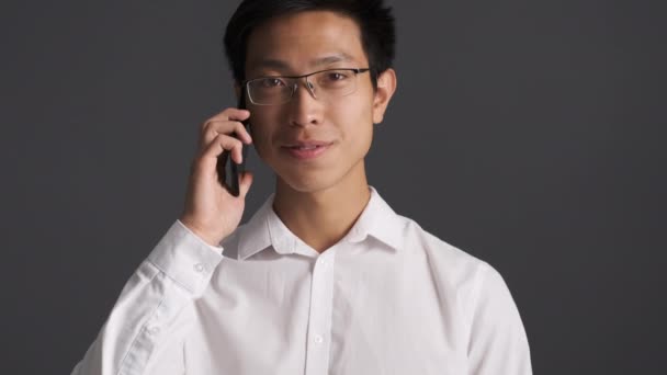 Молодой привлекательный улыбающийся азиат в очках счастливо смотрит в камеру, разговаривая по мобильному телефону на сером фоне
  - Кадры, видео