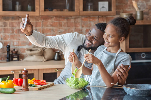 Pai alegre e filha tomando selfie enquanto cozinha na cozinha
 - Foto, Imagem