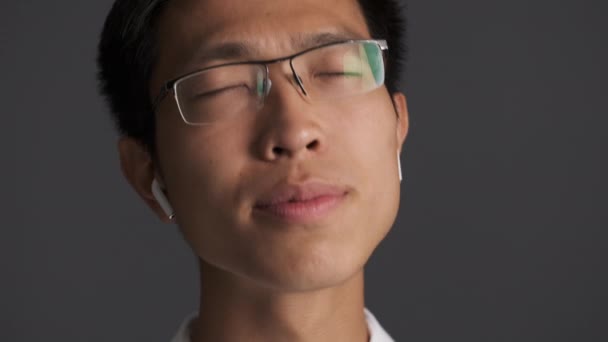 Portret młodego atrakcyjnego Azjaty w okularach słuchającego muzyki w bezprzewodowych słuchawkach i śpiewającego na szarym tle  - Materiał filmowy, wideo