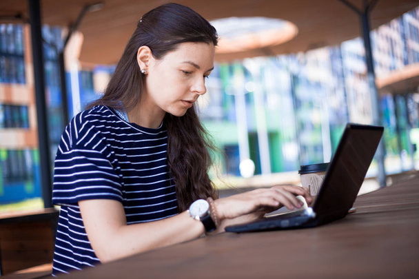 Close-up, portrait of a serious, gently smiling girl student, freelancer, blogger working at a laptop at a wooden table. - Photo, image