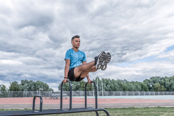 Athlete man handstand summer city on morning exercise, press exercise, twisting legs crossbar, grass clouds background. Active youth lifestyle, fitness workout man. Free space for motivation text. - Foto, afbeelding