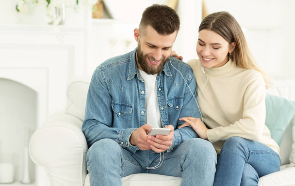 Happy couple sharing music from smartphone sitting on couch - Photo, Image
