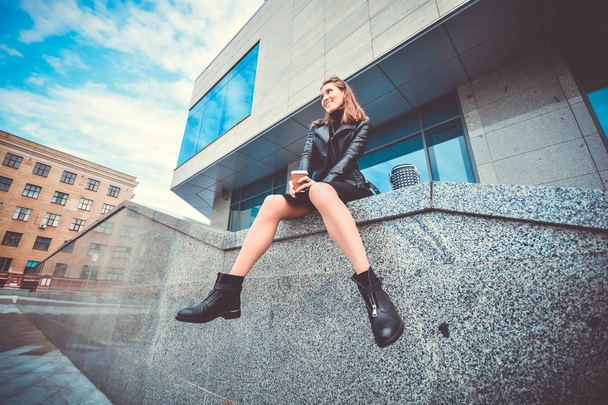 Pretty adult girl in leather boots and jacket sitting on the street and drinking coffee - Foto, Imagem