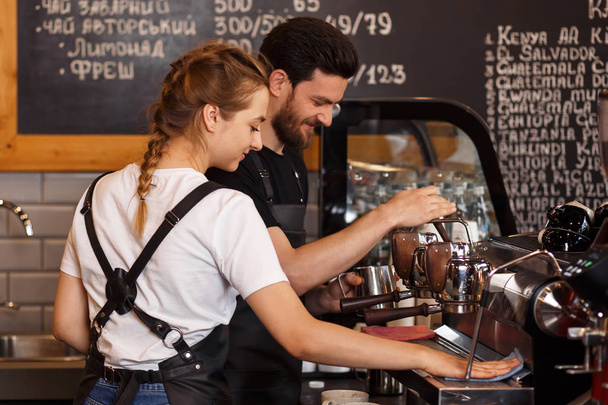 İşteki gülümseyen iki genç barista. Kahve makinesiyle kahve yapan profesyonel bir barista takımı. Mutlu genç adam ve kadın kendi kahve işini geliştiriyorlar. Kahve dükkanı konsepti. - Fotoğraf, Görsel