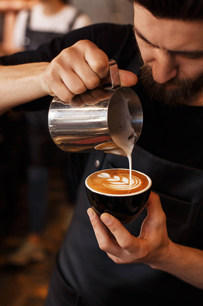 Young smiling male barista making cappuccino. Pouring milk for prepare cup of coffee. Latte art. Morning cup of coffee in caf. Brewing coffee.  - Foto, Bild
