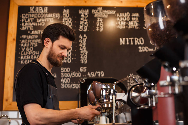 Young smiling male barista making coffee using professional coffee machine. Brewing coffee. Coffee shop concept. - 写真・画像