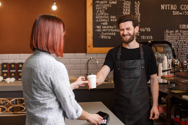 Young smiling male barista gives cup of coffee to customer. Young woman buy morning cup of coffee and paying with contactless credit card. Coffee shop concept. - Фото, зображення