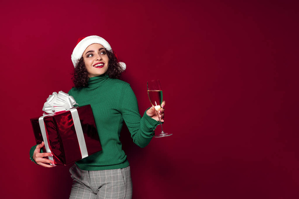 Beautiful smiling woman in santa claus hat, warm green Christmas sweater holding a glass of champagne and gift box on. - Foto, Imagen