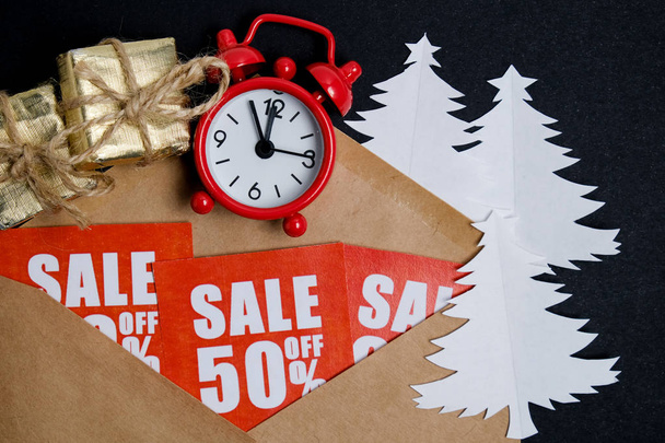 Vintage ρολόι με κουτιά δώρων σε κόκκινες πλάκες με εκπτώσεις με ένα φάκελο τέχνη και χαρτί χριστουγεννιάτικα δέντρα σε μια εφημερίδα τι άνθρωπος σε μαύρο. Χριστουγεννιάτικη ιδέα. - Φωτογραφία, εικόνα