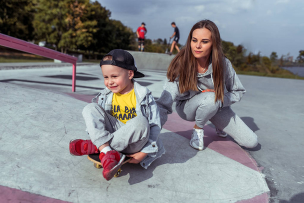 Woman mom rolls playing with child little boy 3-4 years old, autumn summer city, skateboard training, happy smiling having fun, training workout. The boy is riding on board. - Foto, Imagen