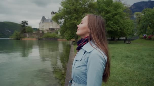 Close up of attractive girl standing in profile on the shores of Lake Annecy. She is wearing blue coat and looking forward with smile. Castle Ruphy is on background. Duingt, France - Záběry, video
