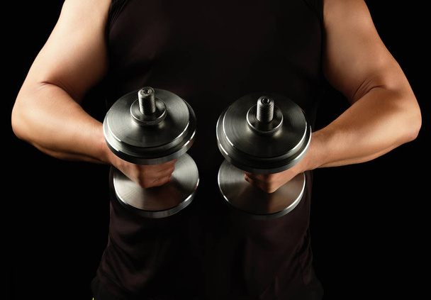 man in black clothes holds steel dumbbells in his hands, his mus - Фото, изображение