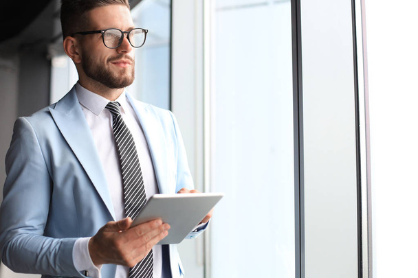 Современный бизнесмен в формальной одежде с помощью цифрового планшета, стоя возле окна в офисе
 - Фото, изображение