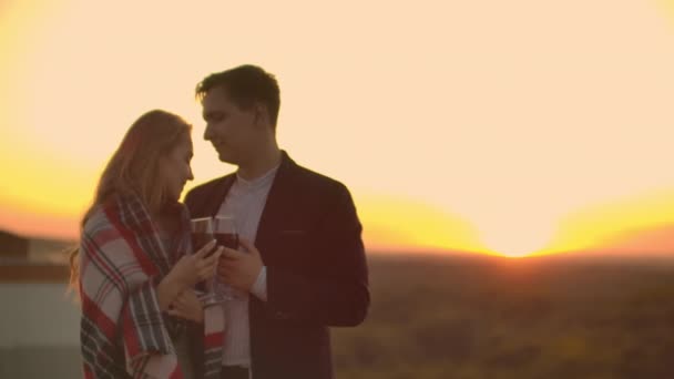 Fiatal házaspár a tetőn átölelve és ivott vörösbort a szemüveg álló öltözött kockás és csodáló a gyönyörű naplemente a város felett. - Felvétel, videó