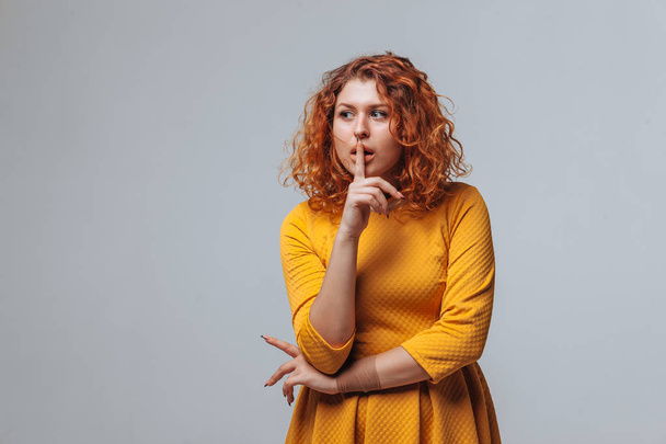 Red-haired girl holds a finger to her lips on a light grey backg - Fotografie, Obrázek