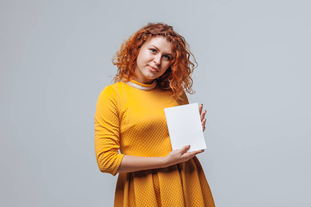 Chica pelirroja sosteniendo un libro blanco sobre un fondo gris claro
. - Foto, Imagen
