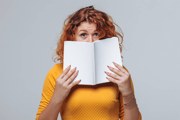 The red-haired girl covered her face with a white book - Фото, изображение