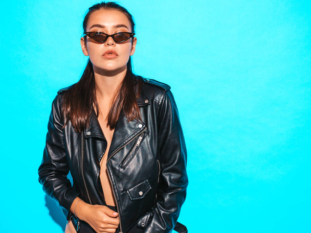 Portrait of young beautiful hipster  bad girl in trendy summer black leather jacket and sunglasses. Sexy carefree woman isolated on blue. Brunette model posing in studio - Foto, Bild