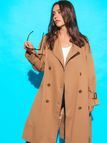 Portrait of beautiful caucasian brunette woman model in brown overcoat and sunglasses.Girl posing in studio near blue wall - Photo, Image