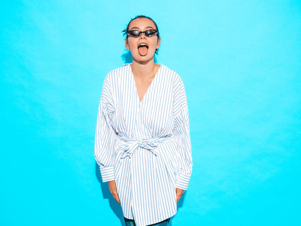 Portrait of young beautiful smiling hipster girl in trendy summer clothes. Sexy carefree woman posing near blue wall. Positive model having fun in sunglasses.Shows tongue - Фото, изображение