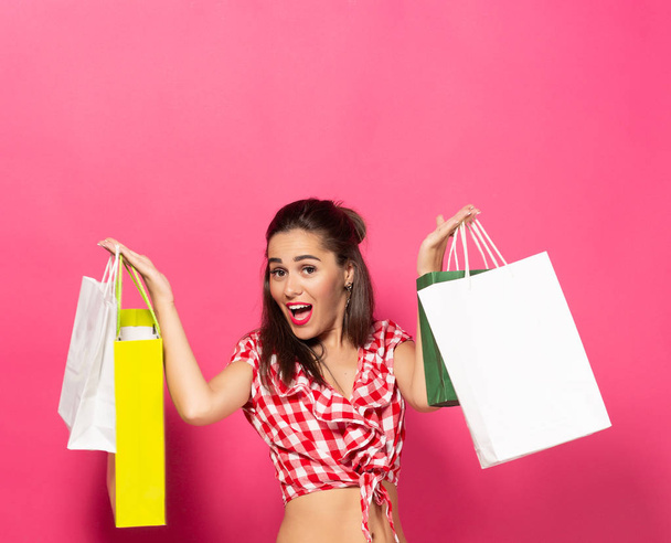 A girl with expressive emotions on her face holds packages with purchases in her hands and looks at the camera. Copy space on pink background - Foto, Imagen