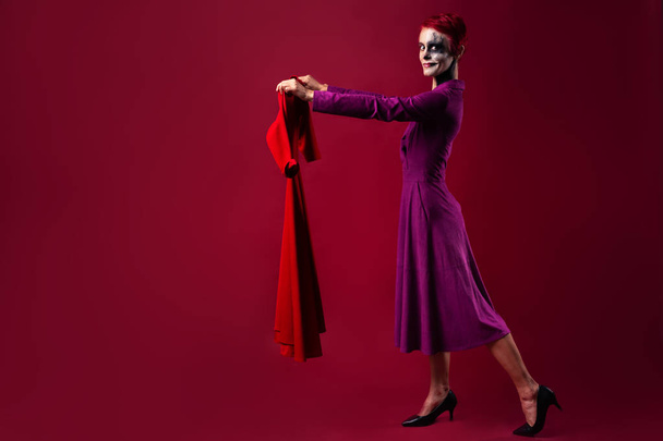 A girl with clown makeup in a purple outfit holds a red dress in her hands. The concept of shopping and choosing clothes - Photo, Image