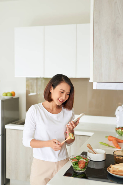 Kaunis nuori nainen yllään esiliina ruoanlaitto terveellistä salaattia keittiössä kotona, käyttäen matkapuhelinta ruoanlaitossa
 - Valokuva, kuva