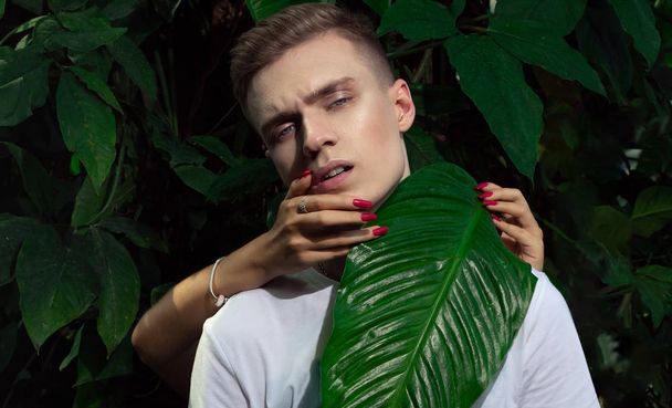 Female hands with red long manicure hold the head of a blond man against a background of green plants. - Photo, image