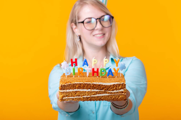 Loca joven alegre en papel sombrero de felicitación celebración tortas feliz cumpleaños de pie sobre un fondo amarillo. Concepto de felicitaciones jubilar
. - Foto, imagen