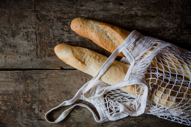 Fresh rosy baguettes in a string bag on a dark wooden retro background. Top view, copy space - Photo, Image