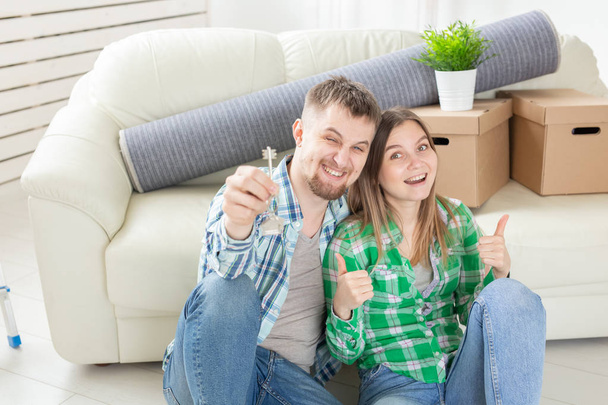 Jovem casal positivo segurando as chaves de um novo apartamento enquanto está em sua sala de estar. Conceito de habitação e hipoteca familiar
. - Foto, Imagem