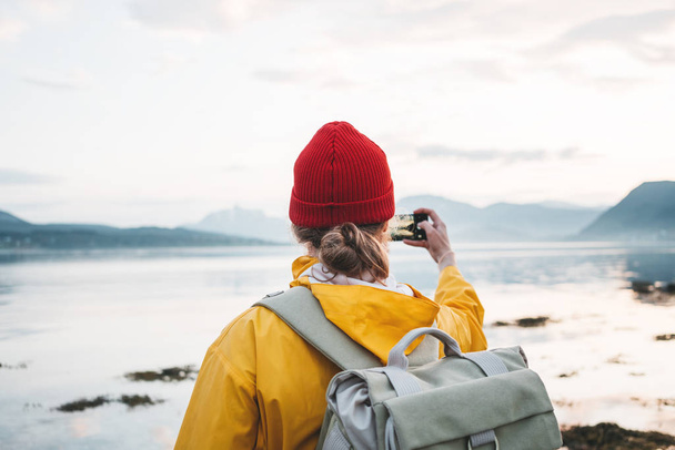 Man traveler wearing yellow raincoat and backpack takes a photo on smartphone of great nordic landscape. Hipster tourist taking photo by smartphone trip summer vacations - Фото, изображение