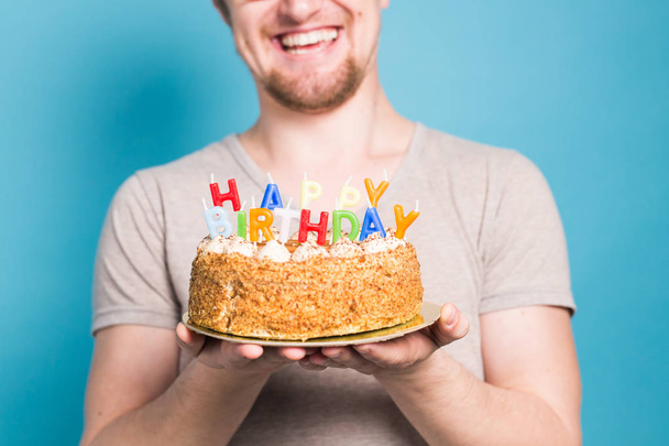 Funny crazy young student in a greeting hat rejoices next to the cake with the inscription happy birthday standing on a blue background. Greetings concept. Copy space - Foto, imagen
