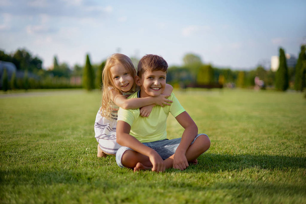 Two happy smiling children posing while sitting on green grass. Cheerful brother and sister laugh together. Happy kids having fun on the park lawn. Children's Day - Zdjęcie, obraz