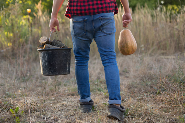 ο άνθρωπος κουβαλάει ένα κουβά με καυσόξυλα και μια κολοκύθα στο φόντο της υπαίθρου - Φωτογραφία, εικόνα