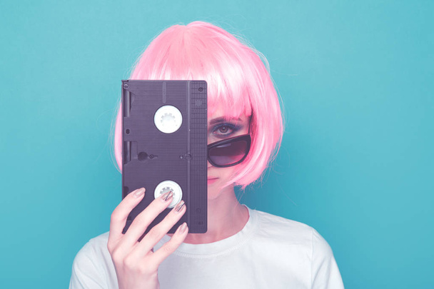 Retro dziewczyna w okularach i różowy peruka trzyma czarną kasetę na niebieskim tle. - Zdjęcie, obraz