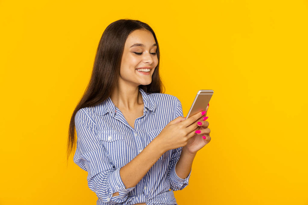 happy female with mobile standing and smiling isolated over the yellow background. - Foto, immagini