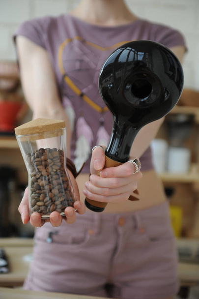 Дівчина тримає в руках керамічний зразок кавоварки та скляної банки зі свіжообсмаженими кавовими зернами
 - Фото, зображення