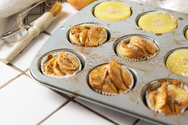 Vdolečky cupcakes s ananasem kruhy a jablečné plátky v muffinovém tácu, na kuchyňské bílé dlaždice v blízkosti s pečivem štětcem. Dort dezert fotografie pro kuchař kniha recept top view - Fotografie, Obrázek