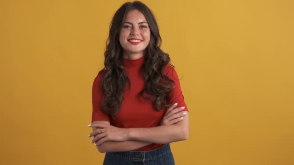 Pretty smiling brunette girl in red top happily looking in camera with hands crossed over yellow background - Footage, Video