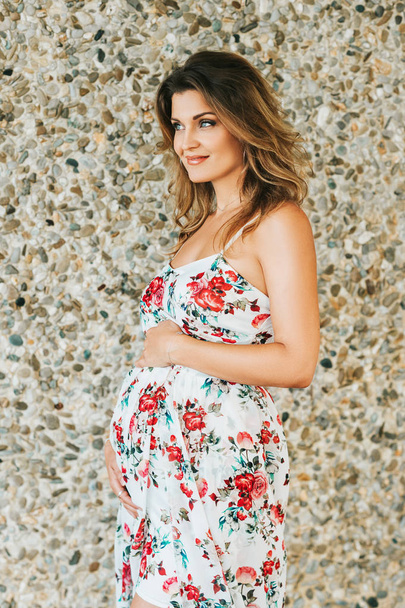 На відкритому повітрі портрет красивої вагітної жінки в квітковій сукні, позує на кам'яному фоні
 - Фото, зображення