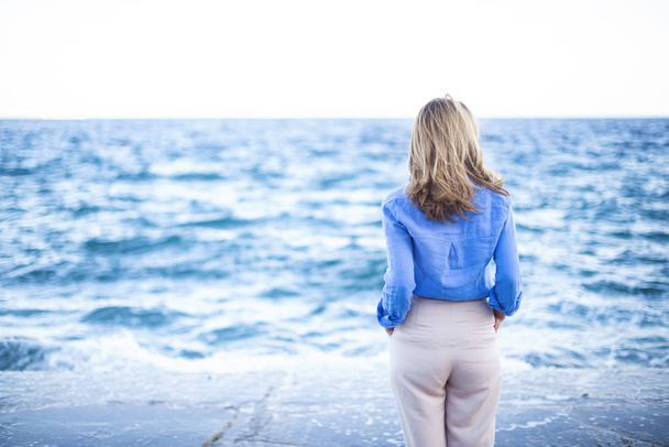 Rear view shot of woman with long hair standing the seaside while looking at waves. - Foto, imagen