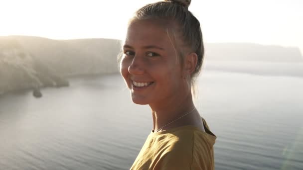 Portrait of a beautiful blonde traveler girl in yellow T shirt on the edge of the cliff, admiring the view and smiling to the camera. Sea view, mountains and sky on the background - Video, Çekim