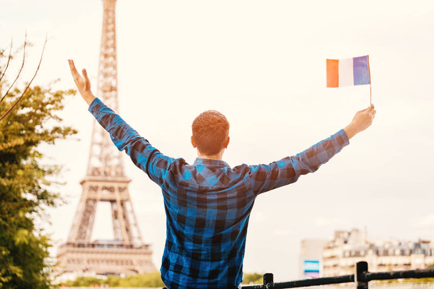 Χαμογελαστός νεαρός που κρατά μια γαλλική σημαία στο φόντο του διάσημου Πύργου του Άιφελ. Ταξίδια, μετανάστευση και σπουδές στην έννοια του Παρισιού - Φωτογραφία, εικόνα