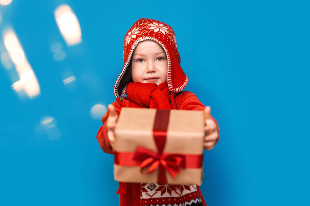 Niño pequeño llevar traje de santa sonrisa y mostrar caja de regalo con lazo rojo sobre fondo azul. enfoque seleccionado. tiempo de Navidad
 - Foto, imagen