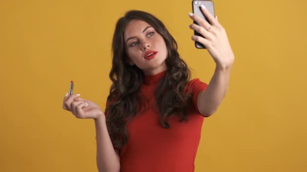 Beautiful smiling brunette girl with red lipstick happily taking selfie on cellphone over yellow background - Filmagem, Vídeo