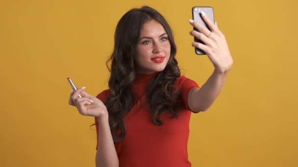 Pretty brunette girl with red lipstick joyfully taking selfie and winking out on cellphone over yellow background - Video