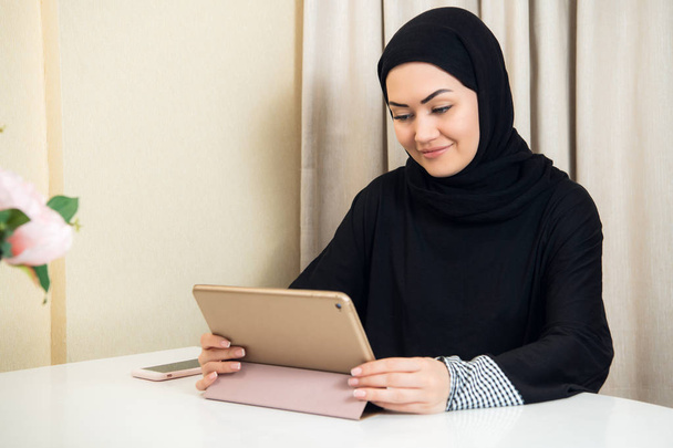 Femme musulmane assise à la maison avec un ordinateur tablette, portant des écouteurs. Se reposer à la maison, se préparer à regarder un film ou écouter une musique
. - Photo, image
