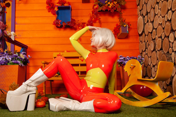 hot pretty blonde woman in bright colorful latex rubber catsuit ready for halloween autumn party and posing near her orange house - Фото, изображение